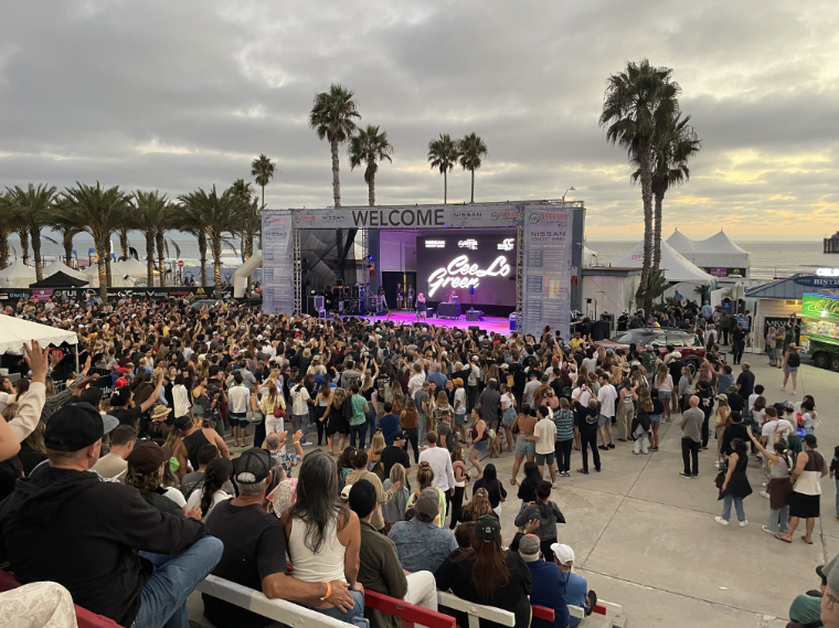 Aerial view of the crowd watching CeeLo Green perform on the Super Girl Surf Pro main stage with palm trees and ocean backdrop.